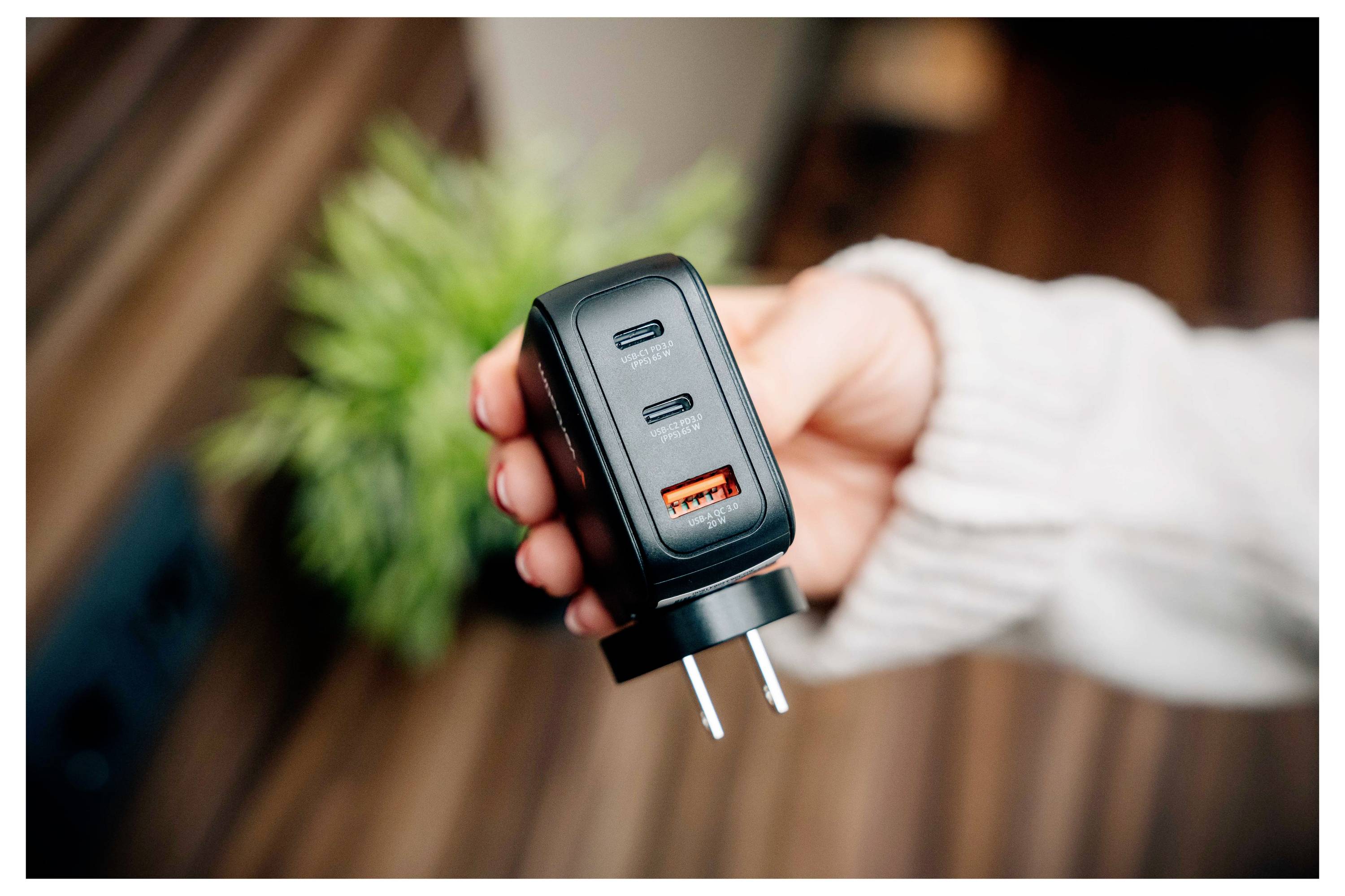 Een hand houdt een zwarte wandlader vast met twee USB-C-poorten en één USB-A-poort, met een onscherpe groene plant en houten oppervlak op de achtergrond.