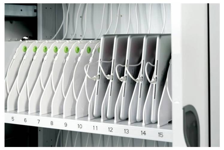 Een rij witte apparaten genummerd van 1 tot 15, elk oplaadbaar in een rek met snoeren. De apparaten hebben groene indicatoren bovenop.