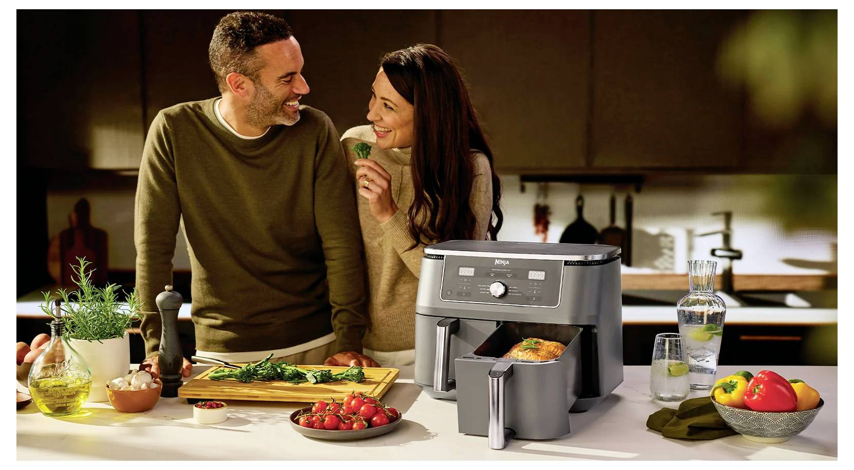 Een stel glimlacht terwijl zij samen in een keuken koken met een airfryer, groenten en brood bereiden, waardoor een warme, gezellige sfeer ontstaat.