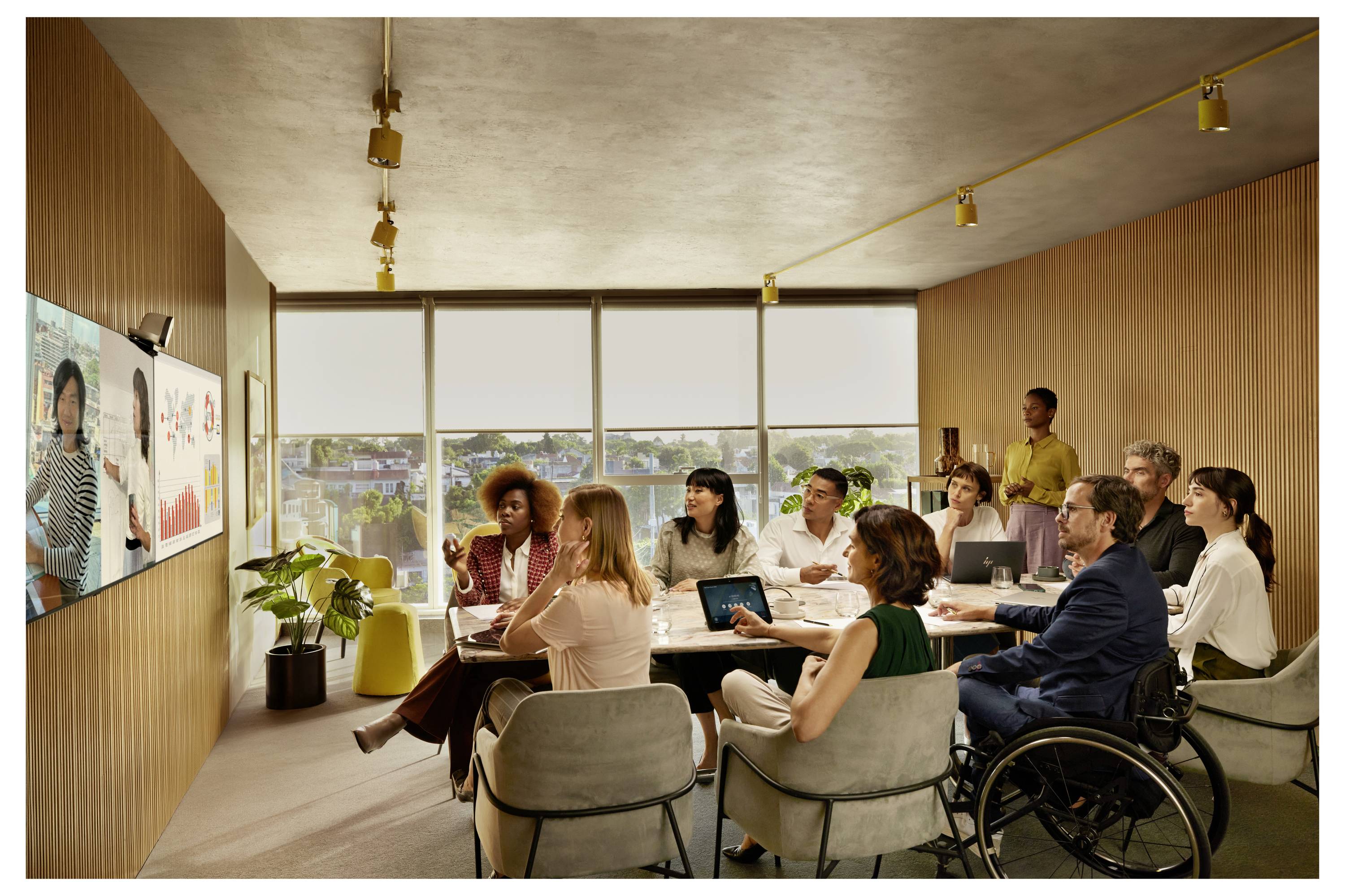 Kantoorbijeenkomst met diverse collega's zittend rond een tafel die naar een presentatie op een wandscherm in een helder conferentieruimte kijken; één deelnemer gebruikt een rolstoel.