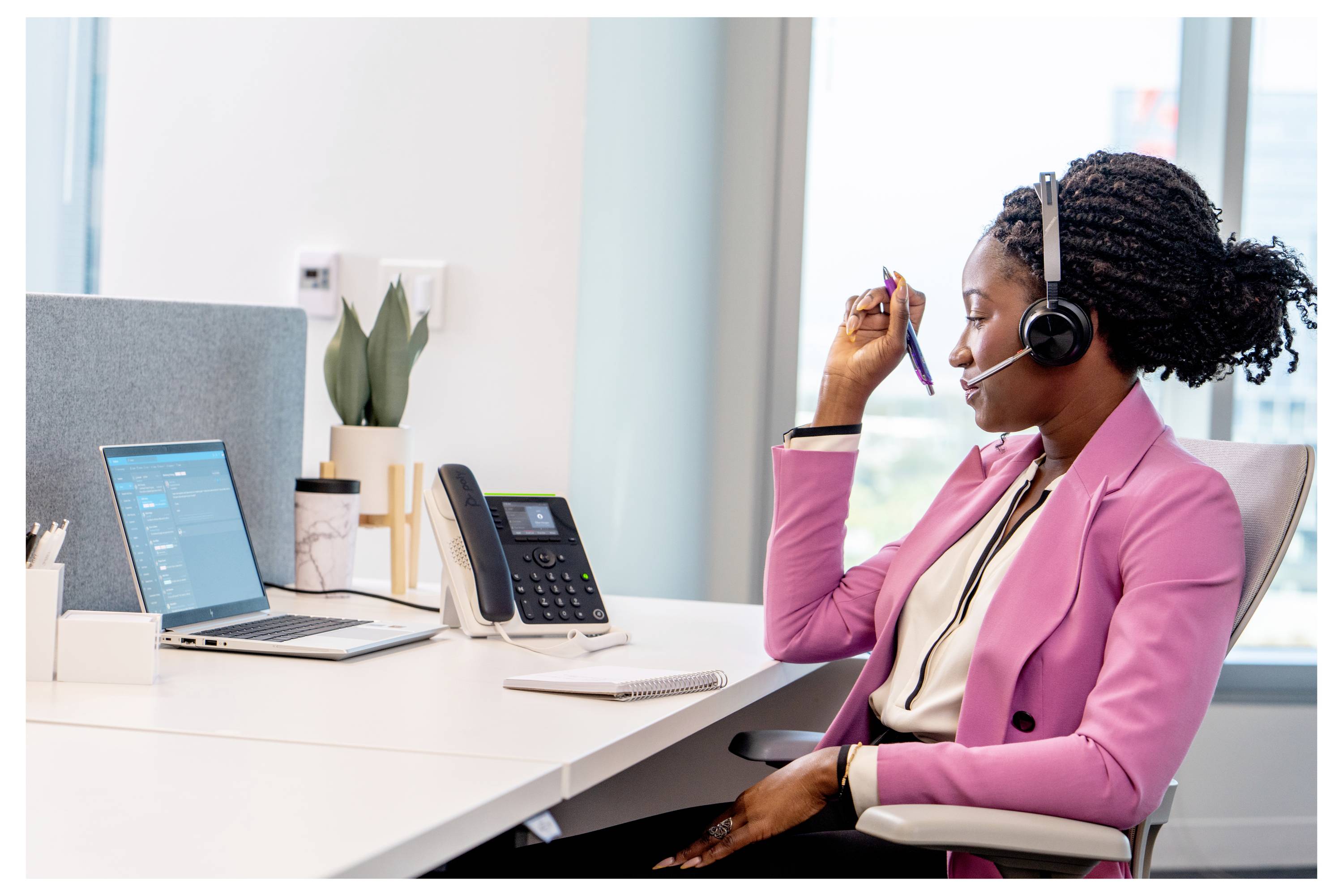 Vrouw die een headset draagt zit aan een bureau op een kantoor, sprekend in een microfoon terwijl ze aan een laptop en telefoon werkt, wat suggereert dat ze een klantenservicegesprek voert.