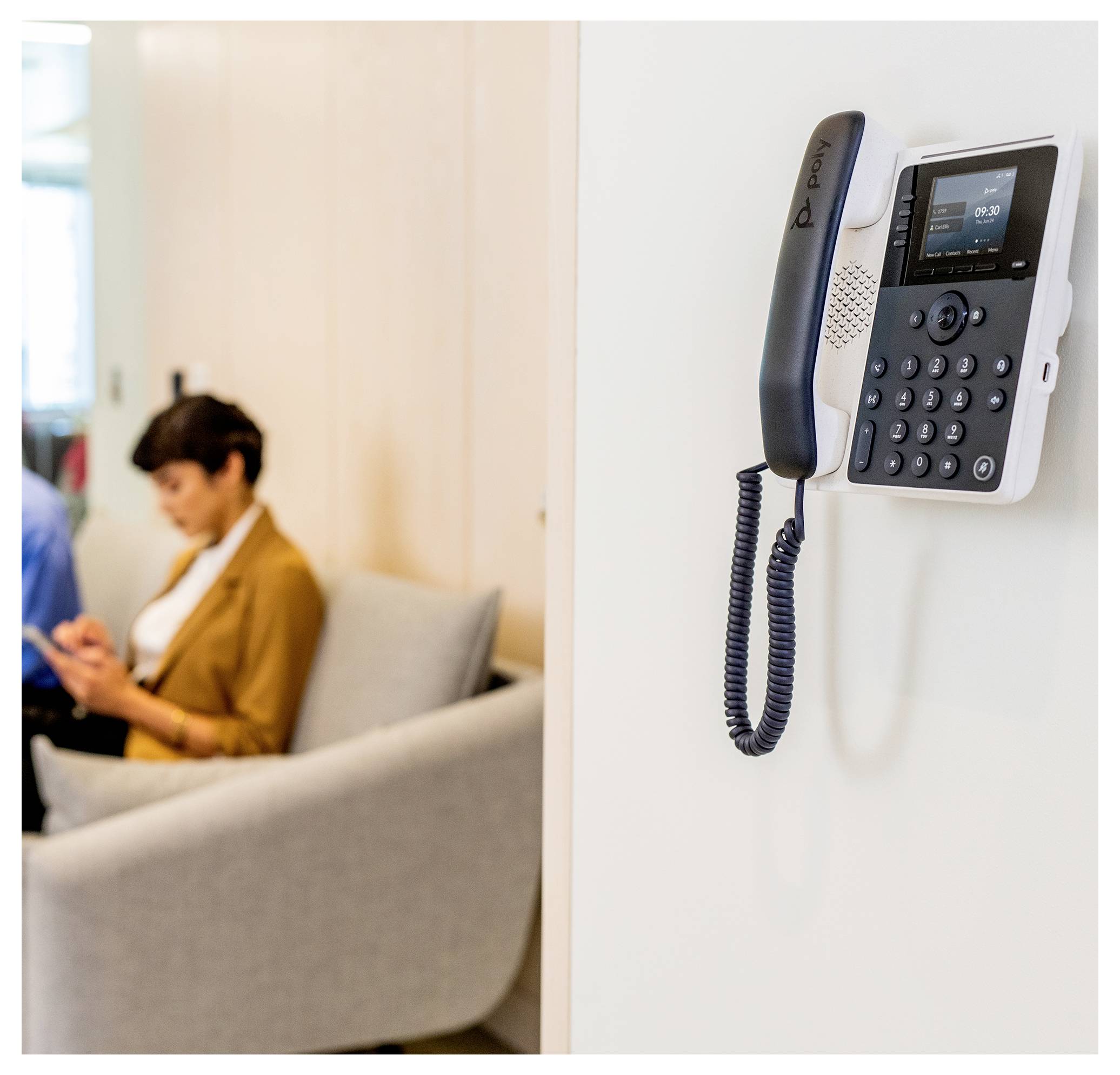 Aan de muur bevestigde kantoortelefoon op de voorgrond; wazig persoon zit op de bank en gebruikt een telefoon op de achtergrond.