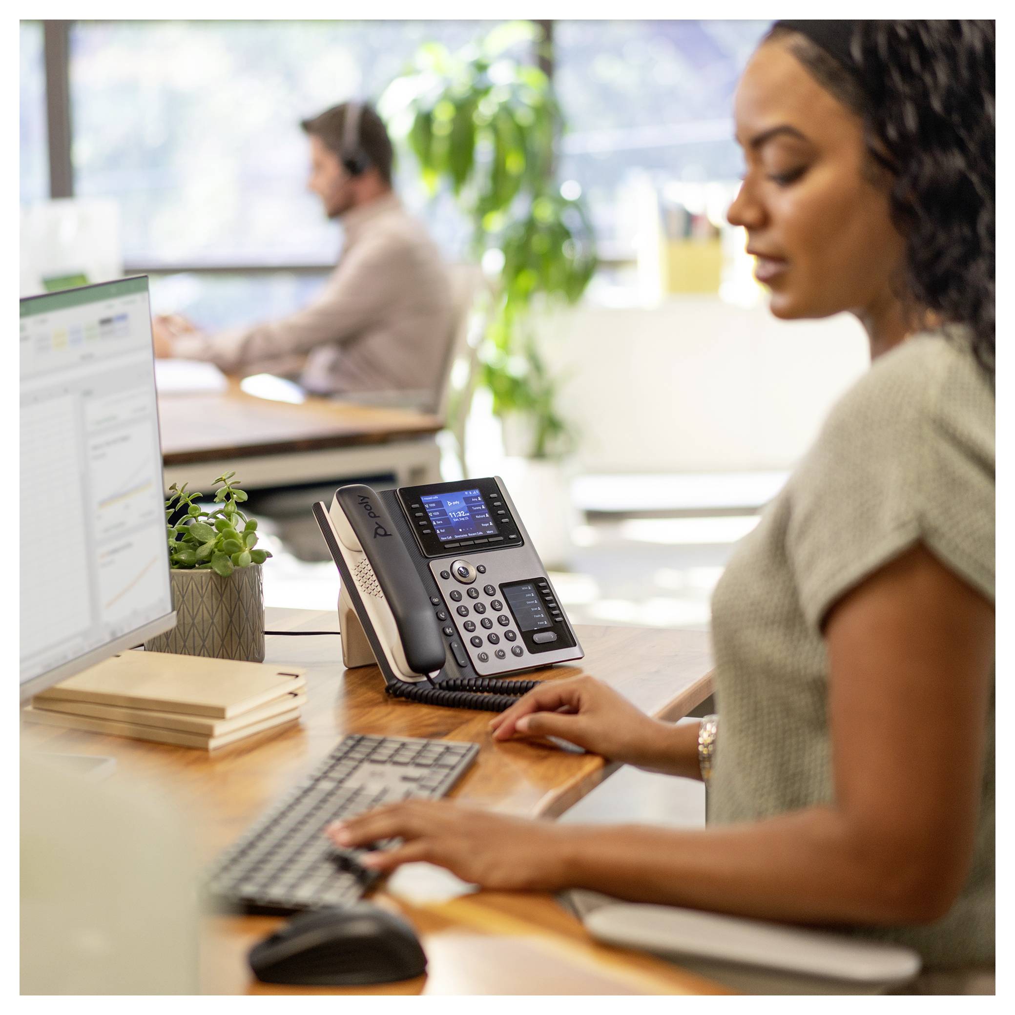 Vrouw die aan een bureau op kantoor typt, een computer en bureautelefoon gebruikt; collega werkt op de achtergrond.