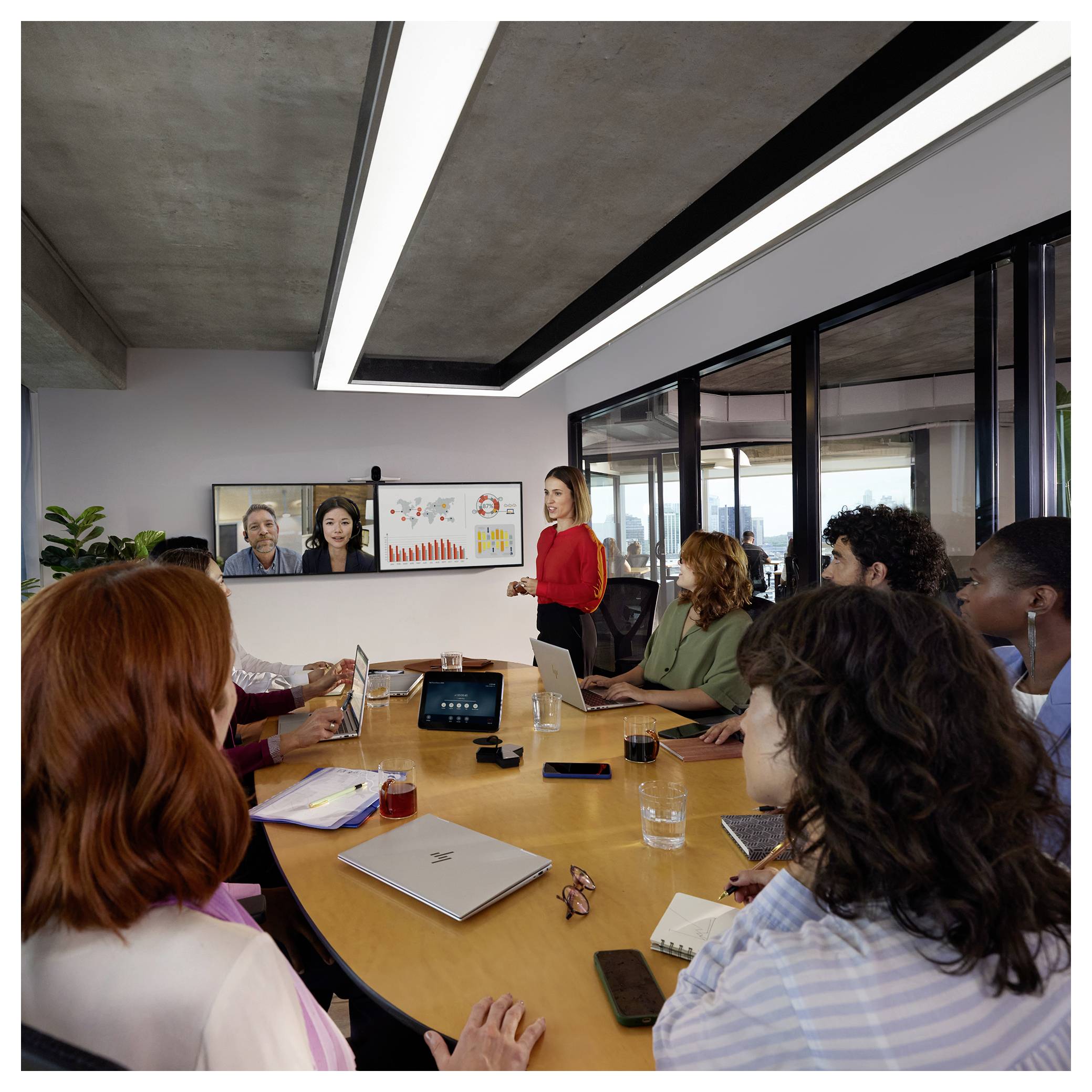Zakenmeeting met een presentator in het rood die tot collega's rond een conferentietafel spreekt, met twee deelnemers op afstand op een scherm en weergegeven grafieken.
