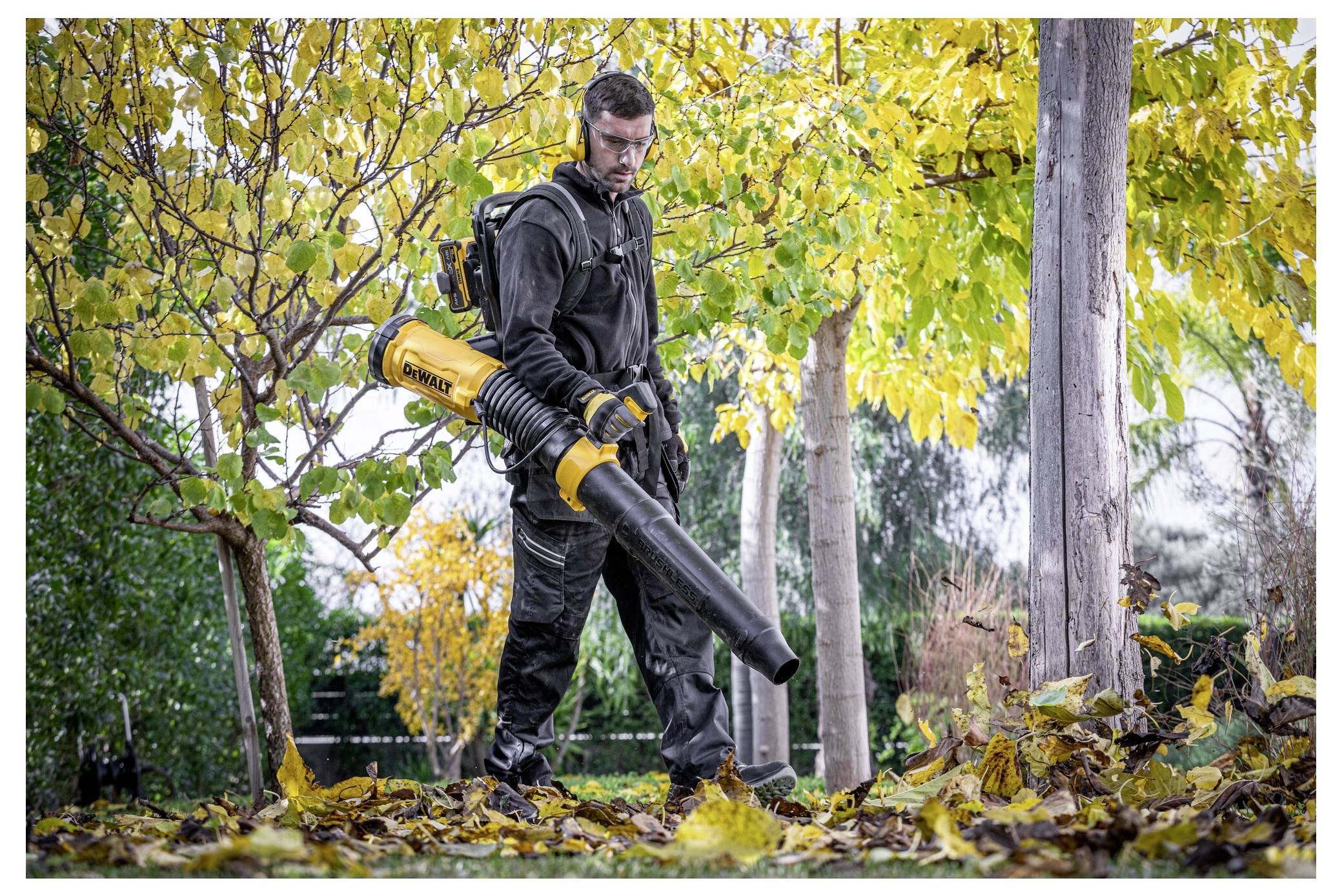Een persoon in werkkleding gebruikt een bladblazer om gevallen bladeren onder een boom met geel gebladerte in een tuinomgeving op te ruimen.