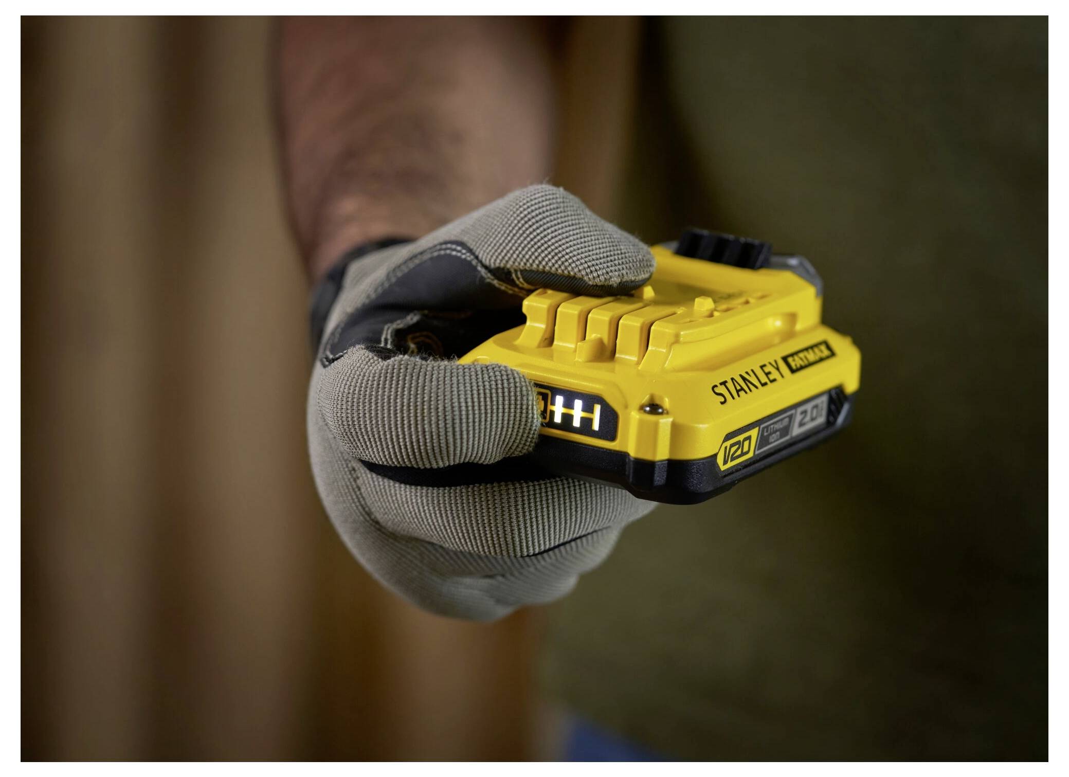 Een gehandschoende hand houdt een gele Stanley FatMax V20 2.0Ah-accu vast, klaar voor gebruik, tegen een vervaagde achtergrond.