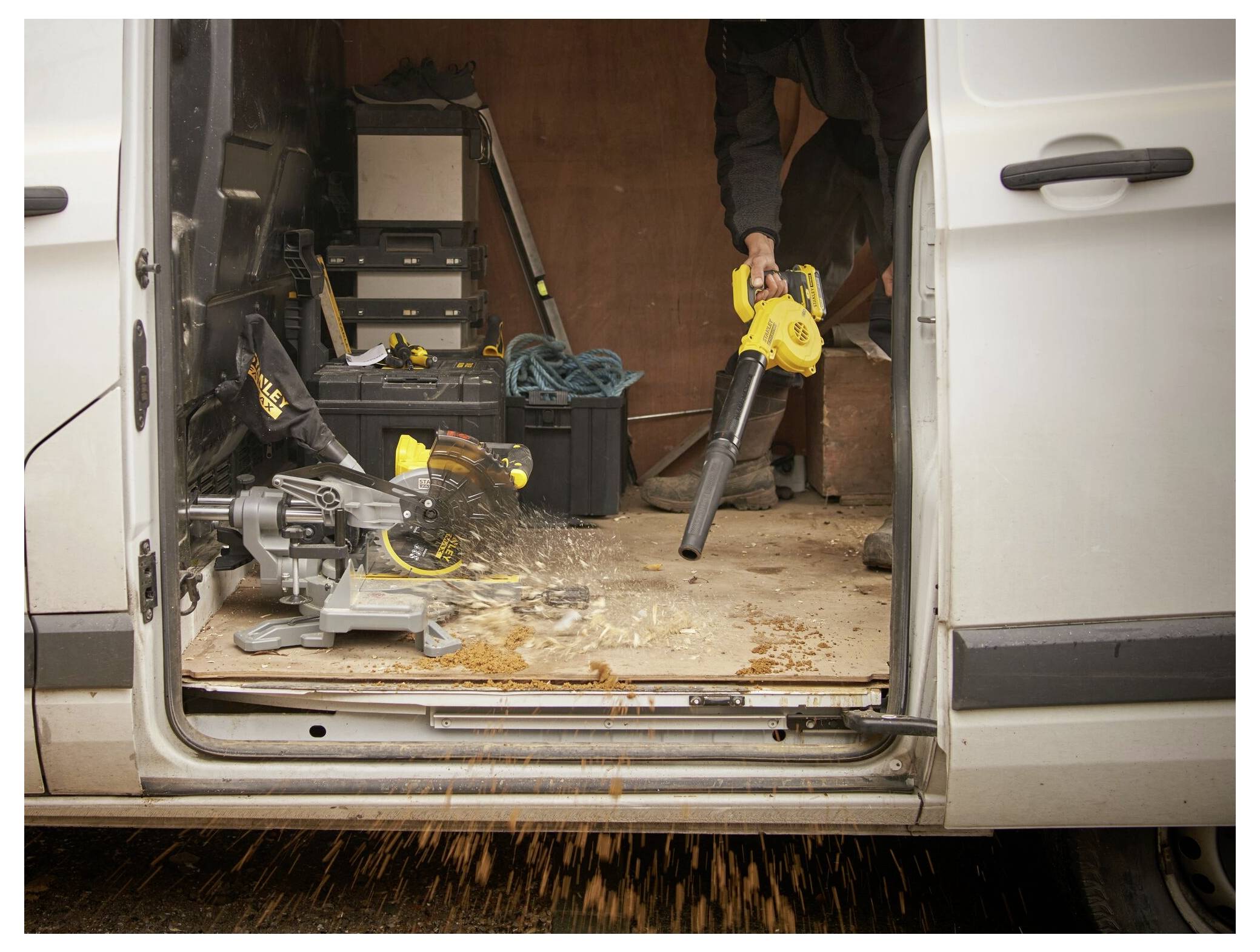 Een persoon gebruikt een bladblazer om zaagsel uit het interieur van een werkbus te verwijderen. De bus bevat verschillende gereedschappen en apparatuur.