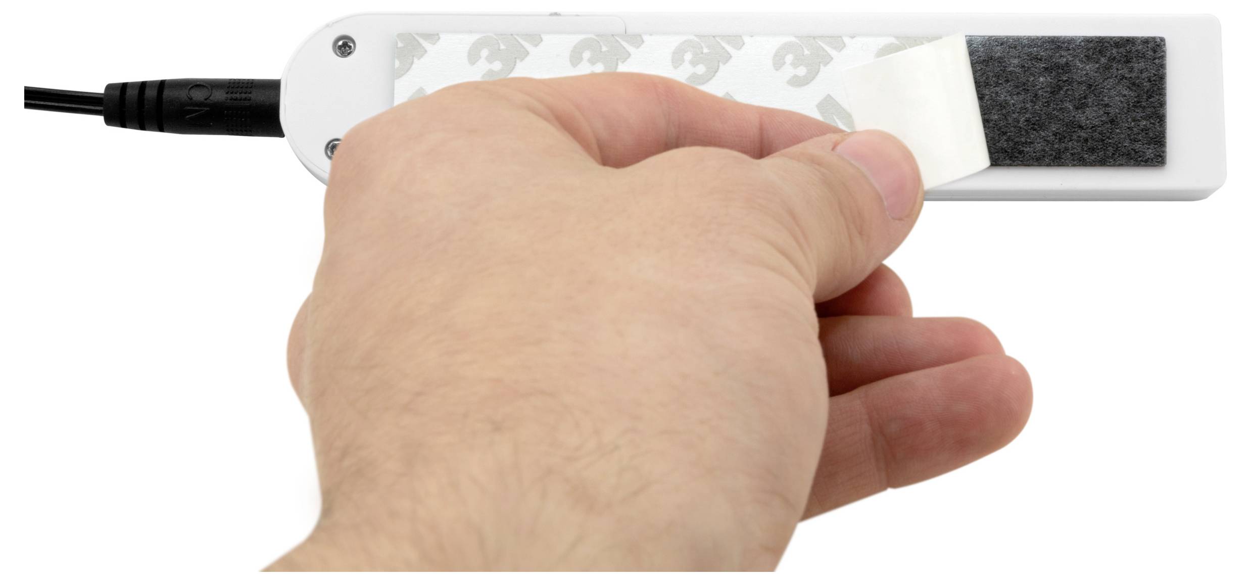 Hand verwijdert een witte kleefstrip van een zwart-wit rechthoekig apparaat met een aangesloten kabel, waarschijnlijk een beschermende achterkant of montagepad verwijderend.