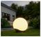 Gloeiende bolvormige buitenlamp op gras in een tuin bij schemering, naast een modern huis en stenen muur.