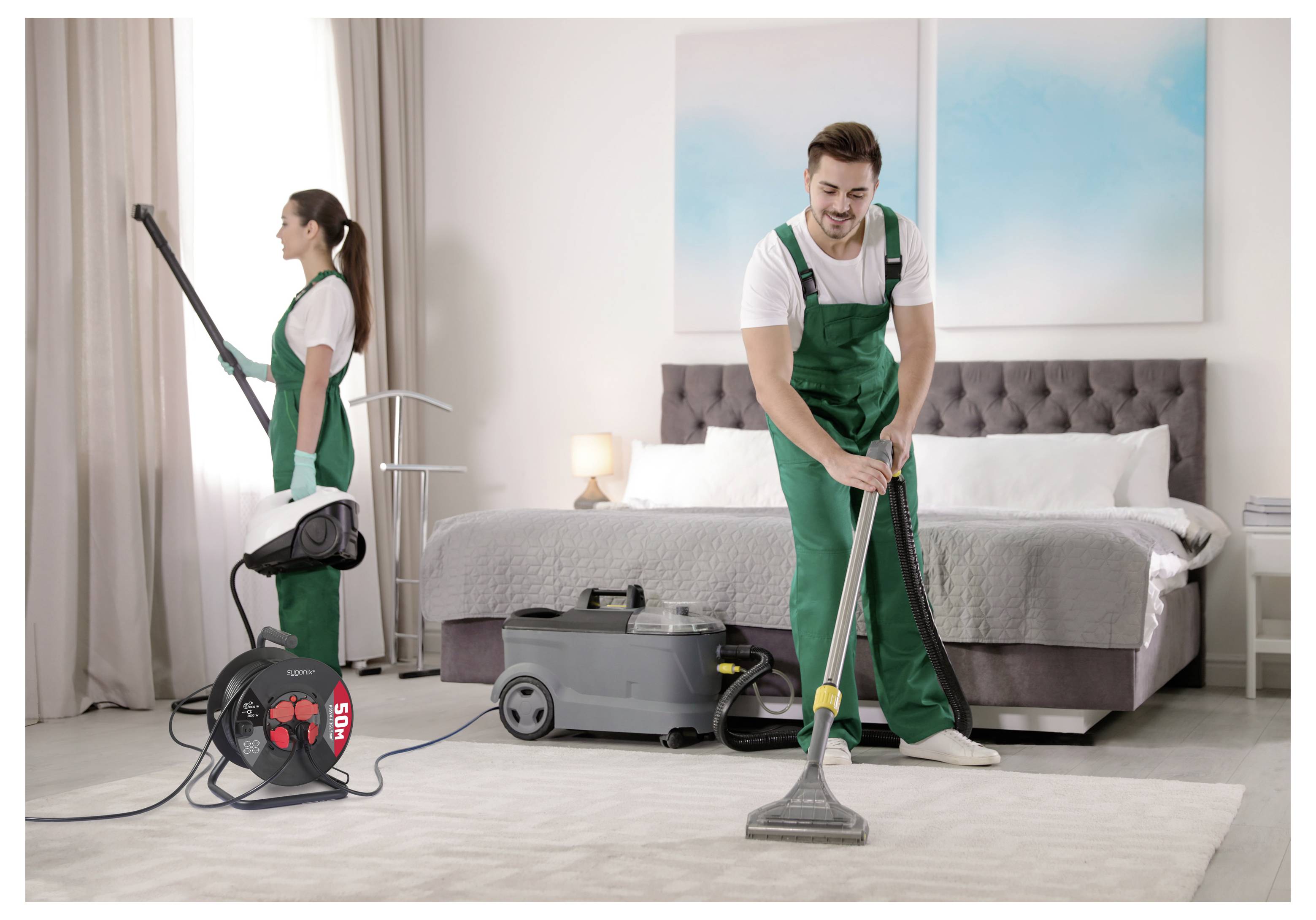 Twee schoonmakers in groene uniformen stofzuigen en stoombmaken een hotelkamer schoon, met apparatuur naast het bed.