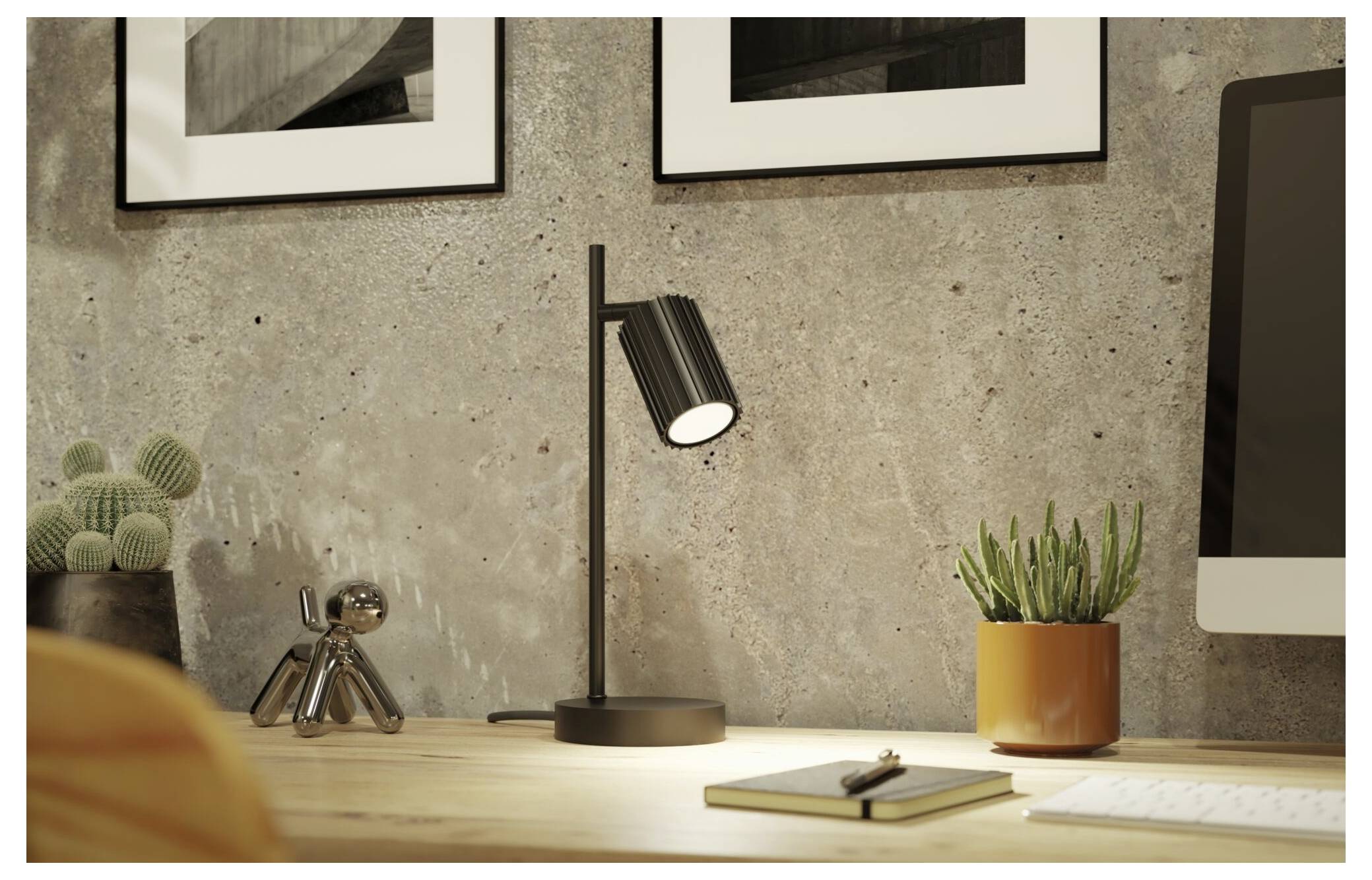 Modern bureauopstelling met verlichte zwarte tafelklamp, potplant, notitieboek en computermonitor tegen een betonnen muur met ingelijste kunst.