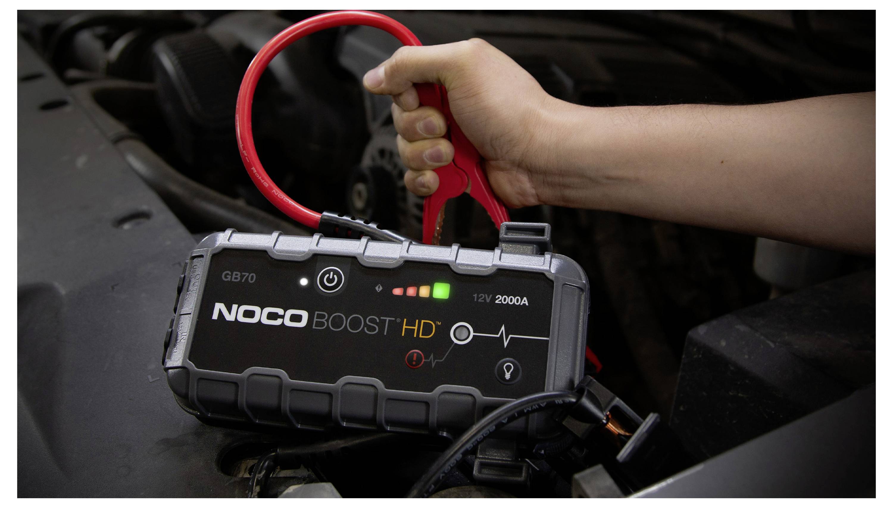 Hand die een NOCO Boost HD draagbare jumpstarter vasthoudt die is verbonden met een autoaccu, met zichtbare accuindicatoren en een 12V 2000A label.