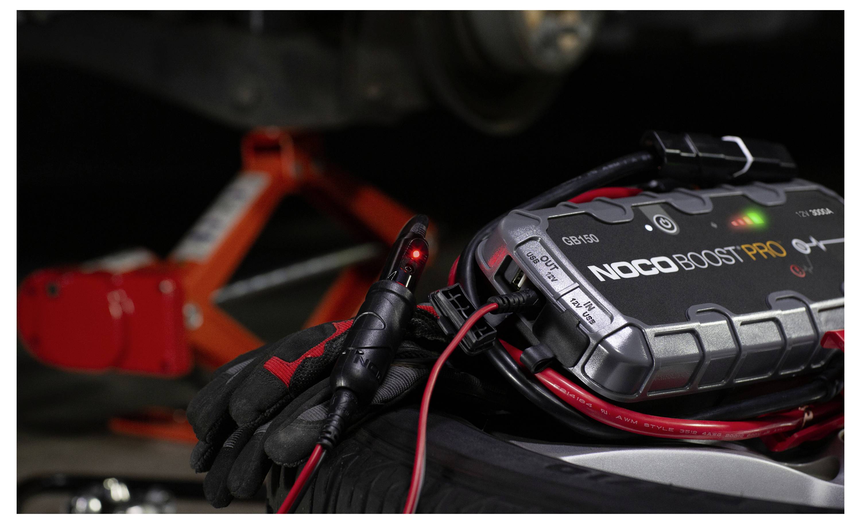 Close-up van een NOCO Boost Pro jumpstarter met kabels en een gehandschoende hand, zittend naast een autoaccu in een motorruimte.