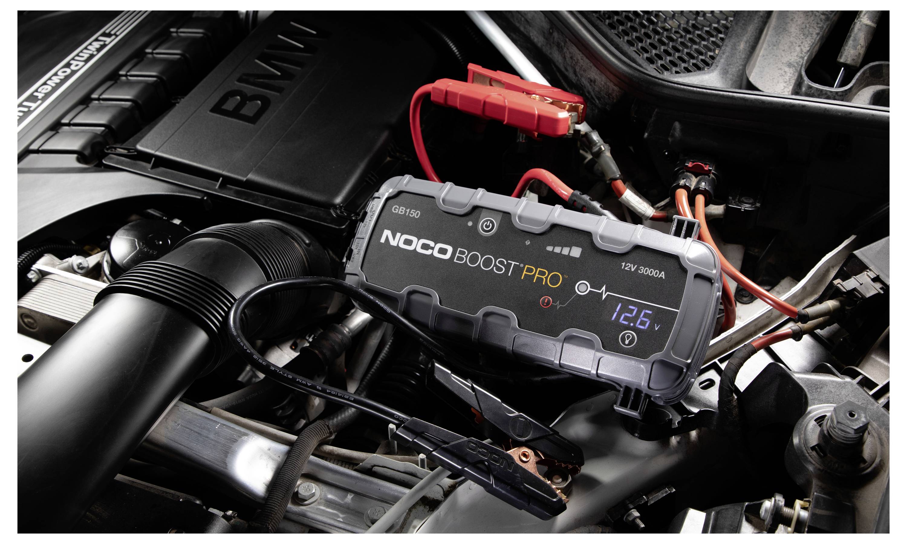 Een NOCO Boost Pro acculader is verbonden met een autoaccu in de motorruimte met rode en zwarte klemmen, waarbij de accuspanning op het display wordt weergegeven.