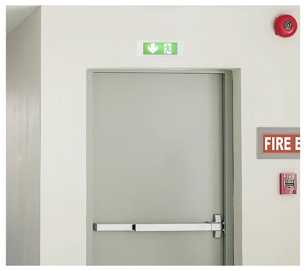 Gesloten grijze nooduitgang in een gang, gemarkeerd met een groen uitgangsbordje erboven en brandveiligheidsequipment ernaast.