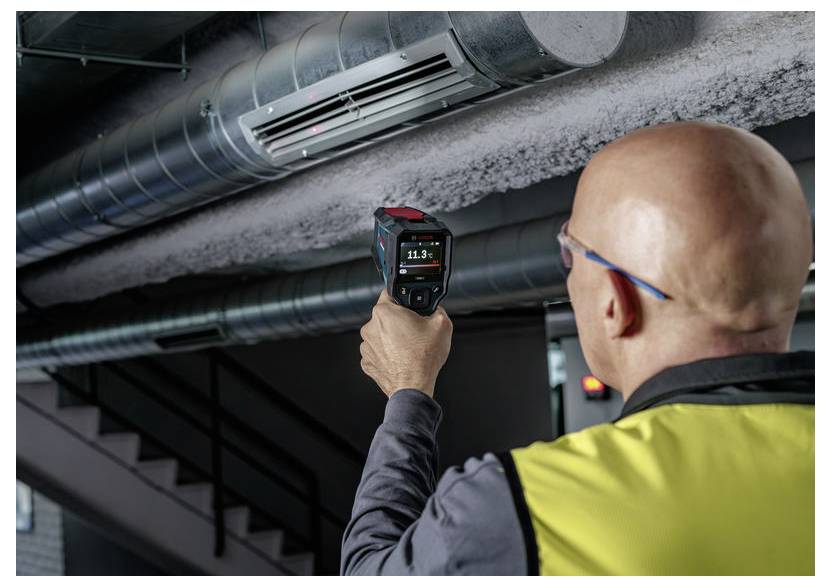 Werknemer in veiligheidsvest gebruikt een draagbare thermische camera om overheadluchtkanalen in een industrieel gebouw te inspecteren.