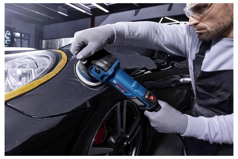 Een nauwkeurige man met handschoenen en veiligheidsbril polijst de voorscherm en koplamp van een auto met een draadloze polijstmachine in een garage.
