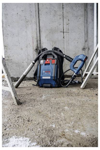 Blue Bosch nat/droog stofzuiger met slang en hulpstukken op een stoffig bouwterrein tegen een betonnen muur.