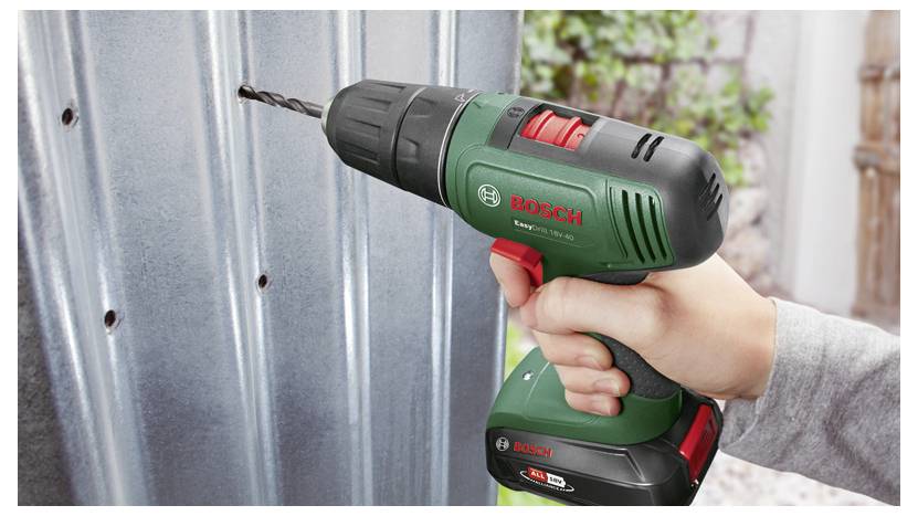 Nahaufnahme van een hand die een Bosch-accuboormachine gebruikt om in een metalen plaat of hekpaneel buiten te boren.