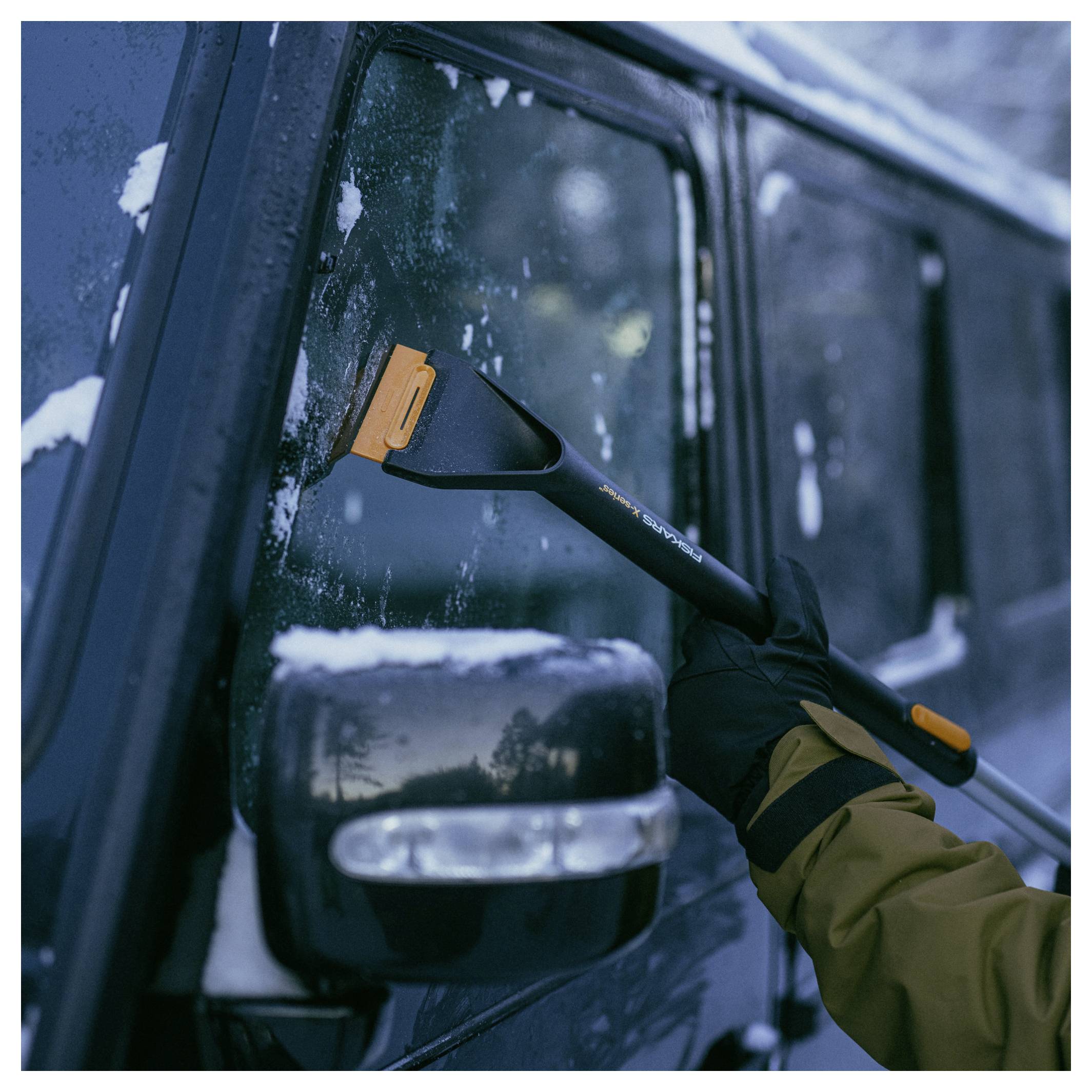 Gehandschoende hand die een sneeuwborstel gebruikt om sneeuw en ijs van een voertuigraam en zijspiegel te schrapen.