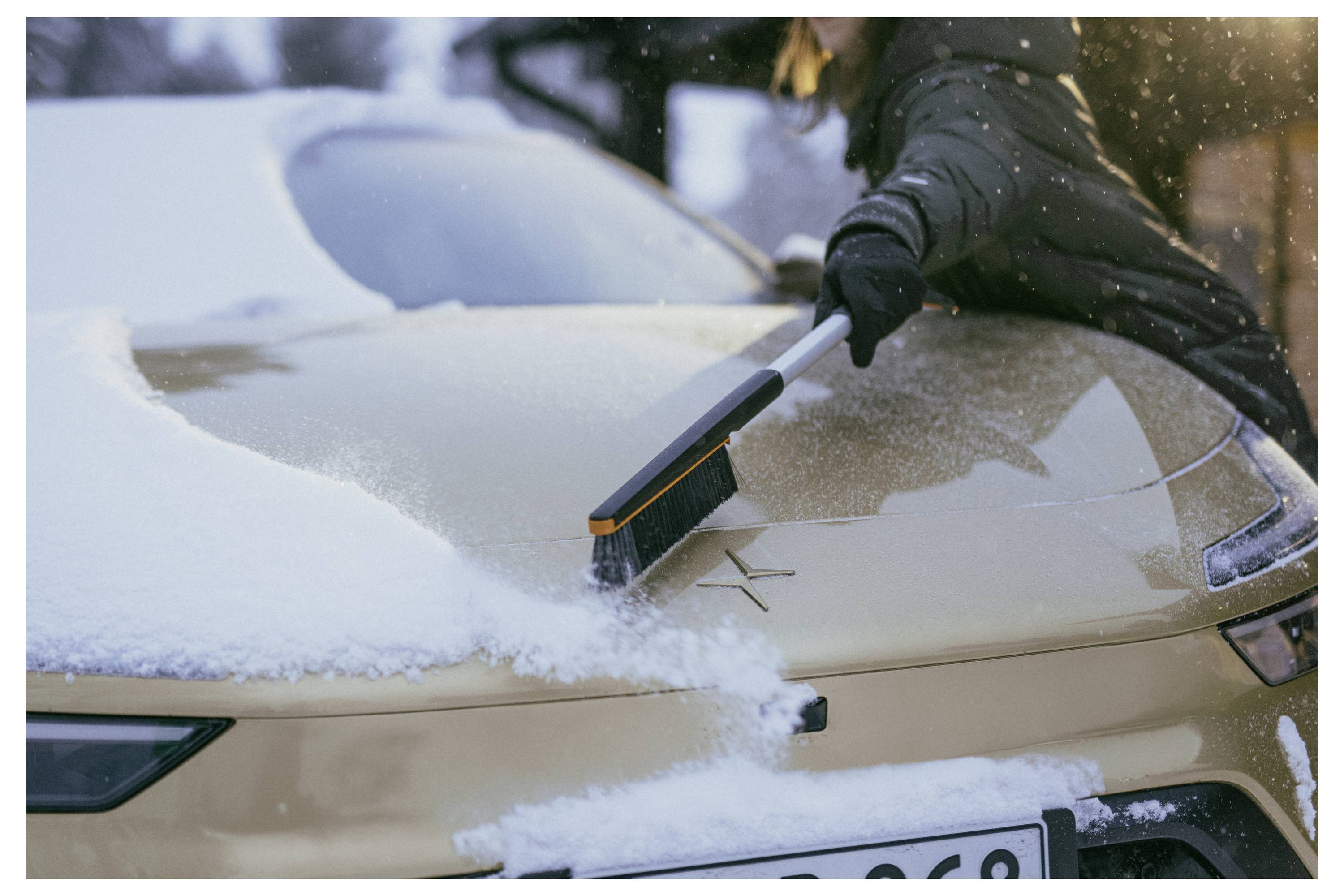 Een persoon veegt sneeuw van de voorruit van een gouden auto met een oranje sneeuwborstel in de winter.