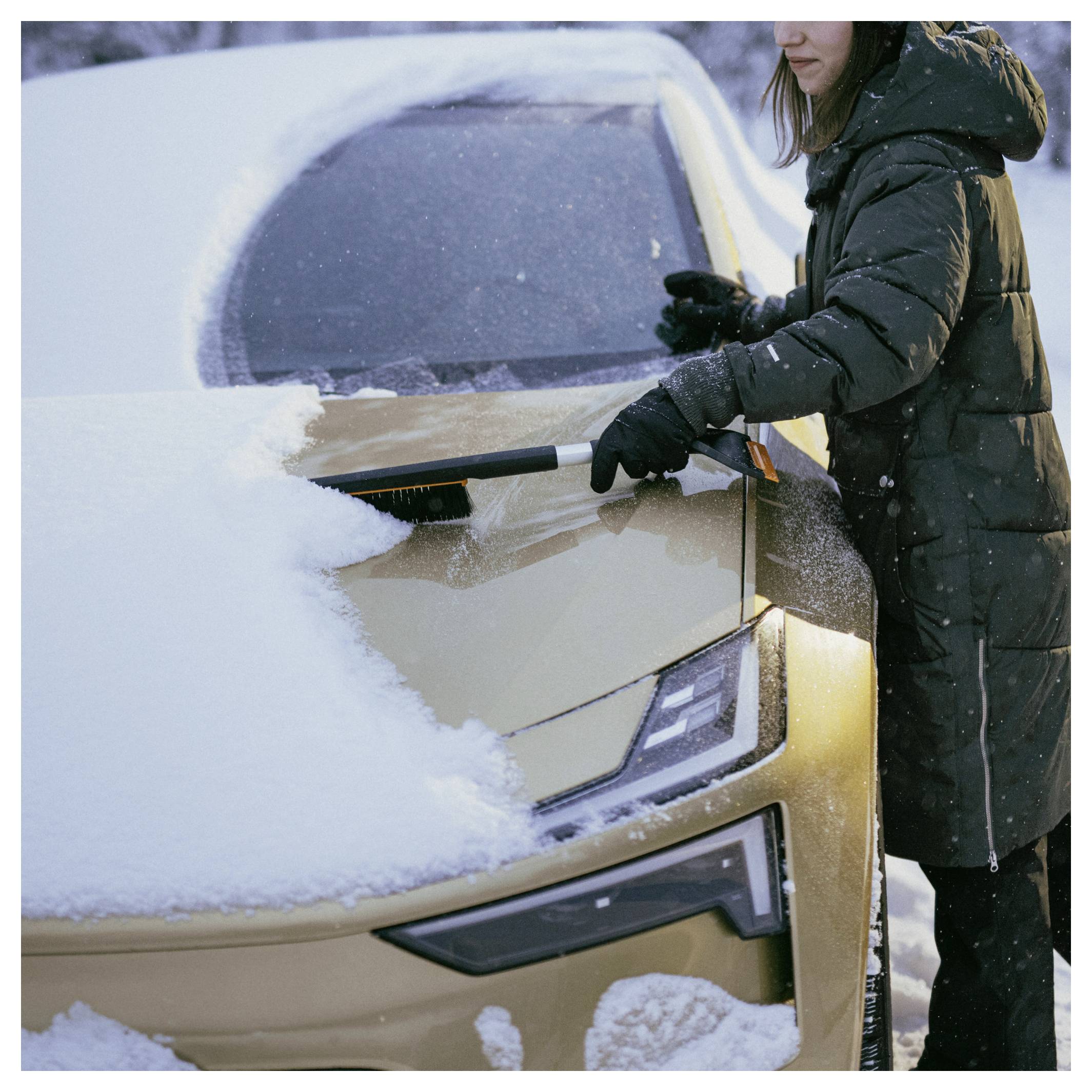 Persoon in winterjas die sneeuw van een autoraam en motorkap veegt in een sneeuwscène.