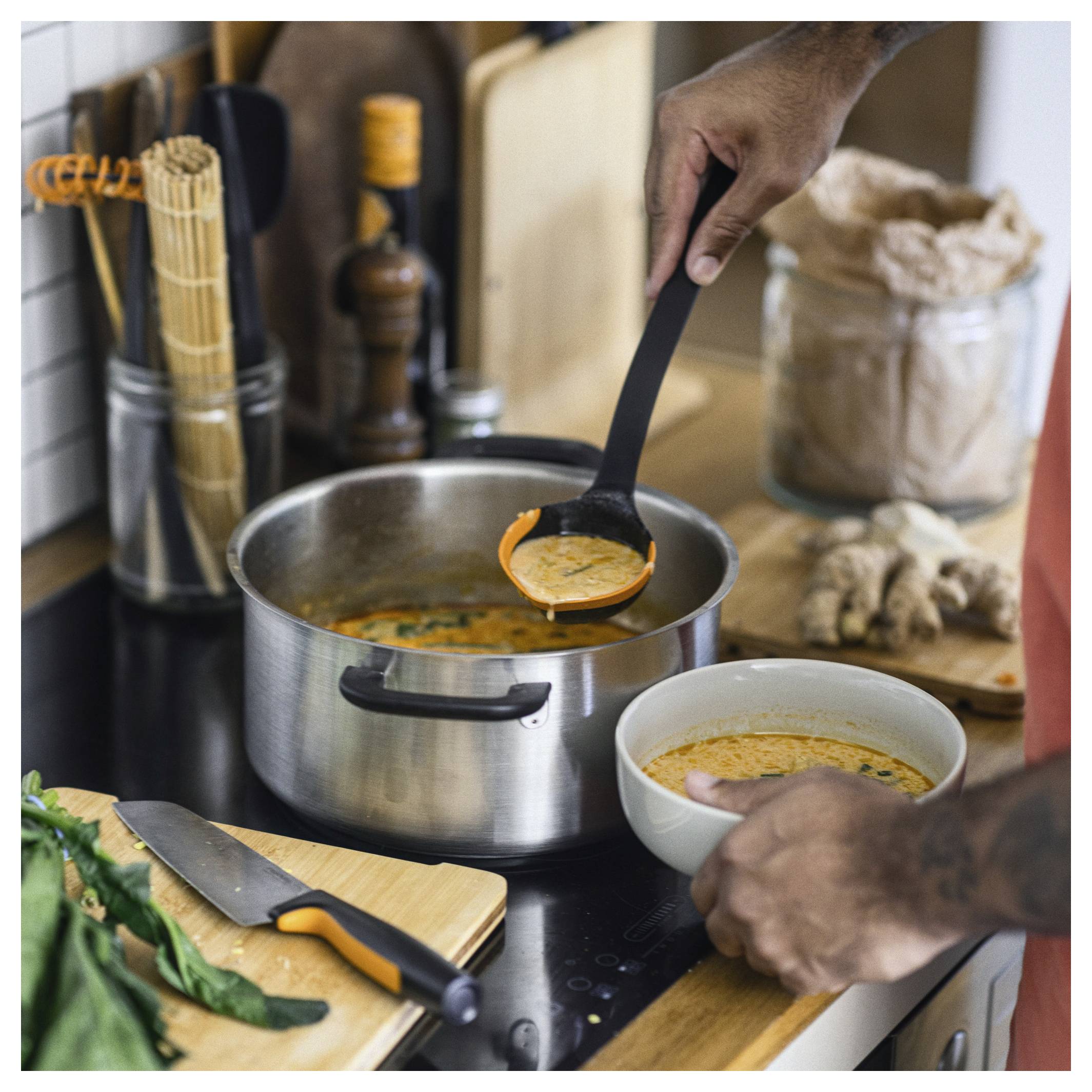 Een persoon scheidt soep van een pot in een kom op een keukenplateau, met keukengerei en ingrediënten in de buurt.