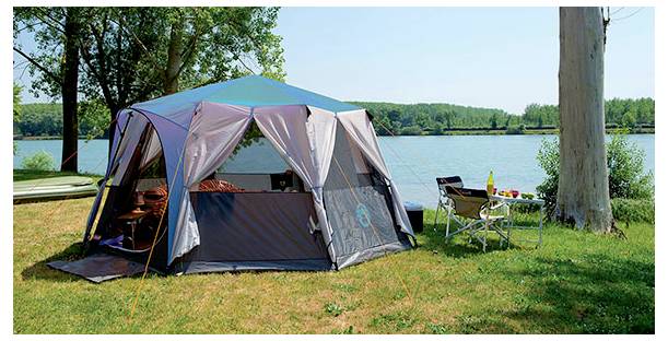 Campingtent opgezet op een graszige oever van een meer, met een tafel en stoelen ernaast en een meer en bomen op de achtergrond.