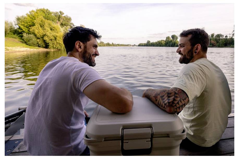 Twee mannen zitten op een bootdek naast een koelbox, glimlachend en pratend terwijl zij over een kalm meer naar de zonsondergang kijken.