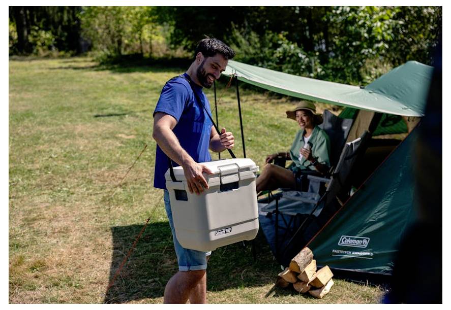 Man die een koelbox draagt naast een kampeerplaats met een groene tent en een zittende kampeerdeder, met gestapeld brandhout op de voorgrond.