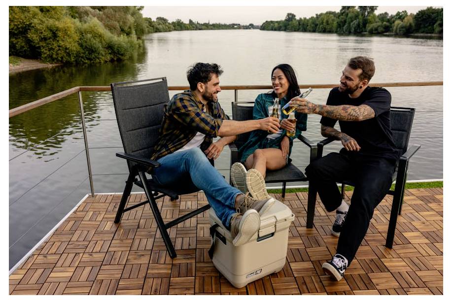 Drie vrienden ontspannen zich op een dek aan de rivier, glimlachend en met hun drankjes toostend terwijl zij in stoelen naast een koelbox zitten.