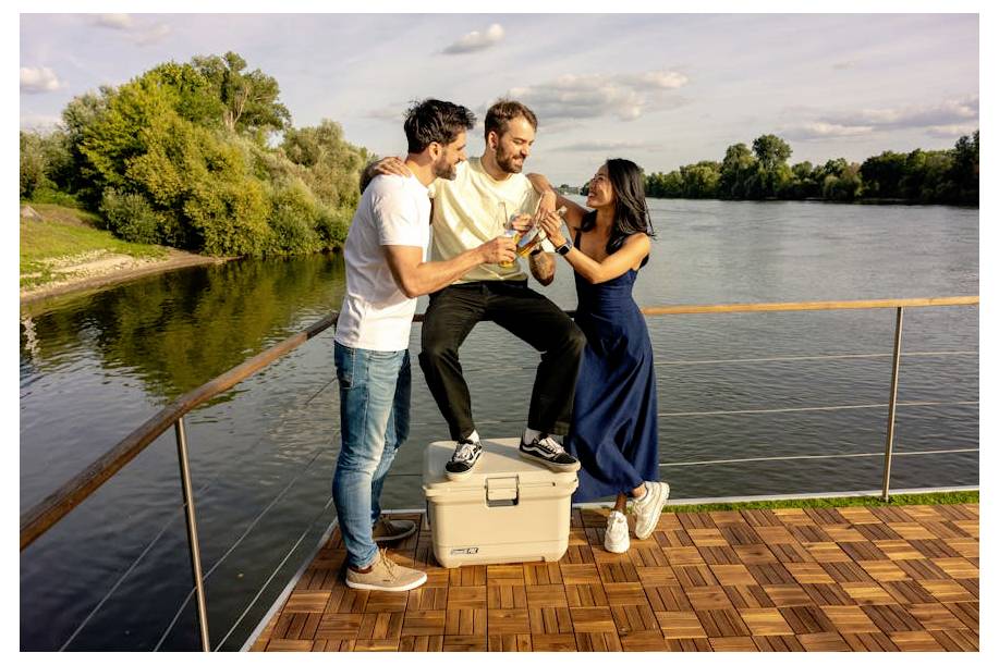 Drie volwassenen op een bootdek delen een drankje aan een rivier bij zonsondergang, glimlachend en samen vierend.