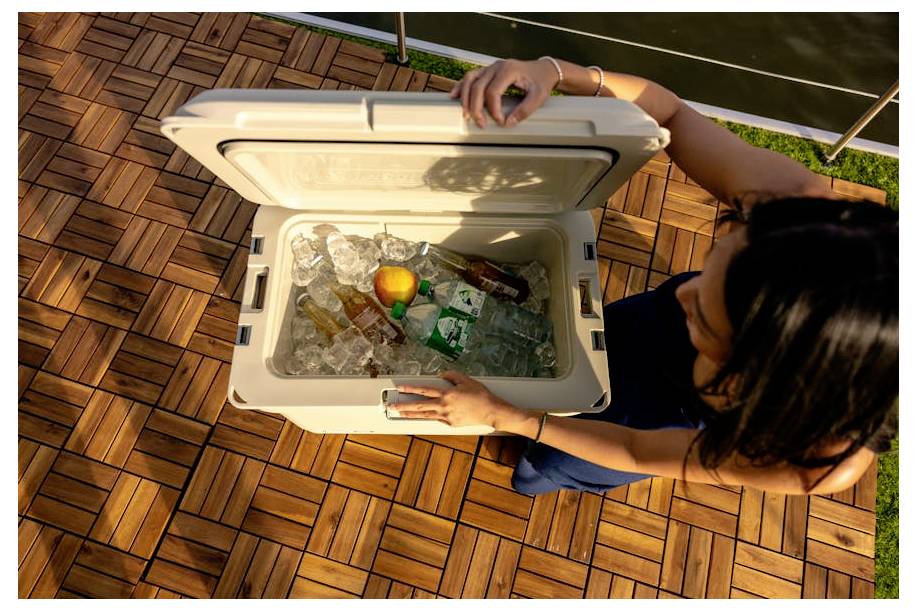 Een vrouw opent een koelbox gevuld met ijs, flessen en een oranje drank, van bovenaf gezien op een houten terras buiten.