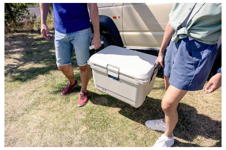 Twee mensen dragen een grote witte koelbox naast een geparkeerde bestelwagen op gras, wat wijst op kamperen of buitenreizen.