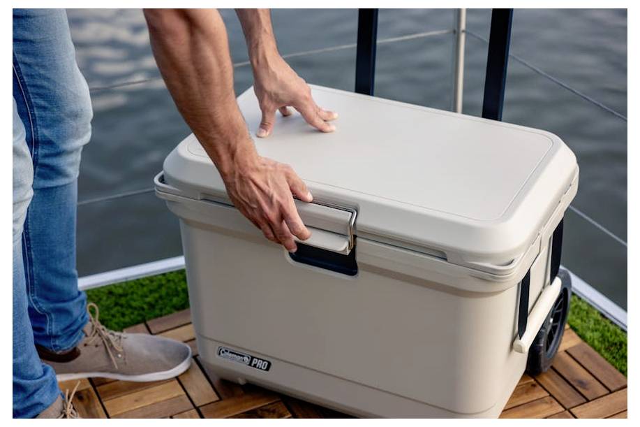 Een persoon sluit een grote witte koelbox op een houten steiger aan het water af, met handen op het deksel en de sluiting.