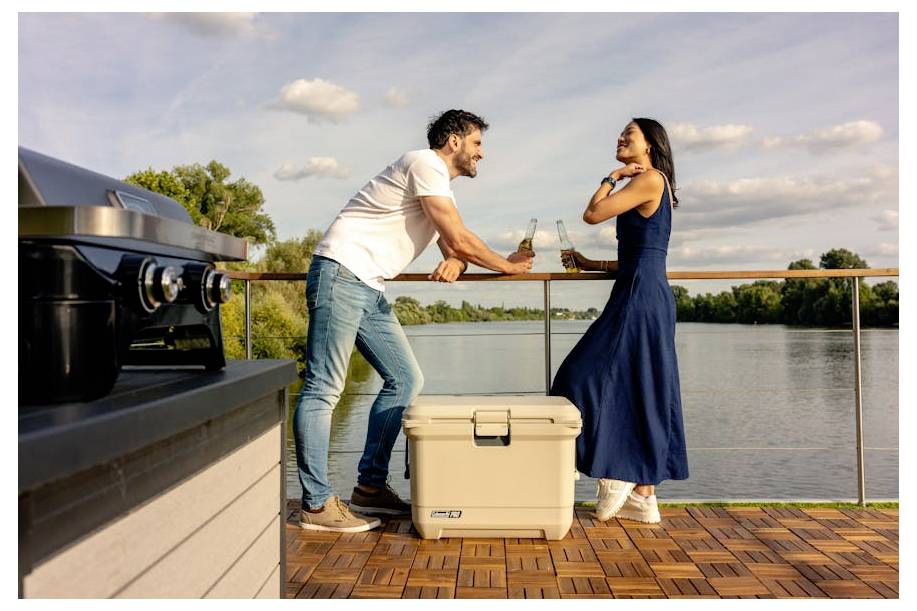Man en vrouw chatten en houden drankjes vast op een terras aan het meer naast een koelbox en grill bij zonsondergang.