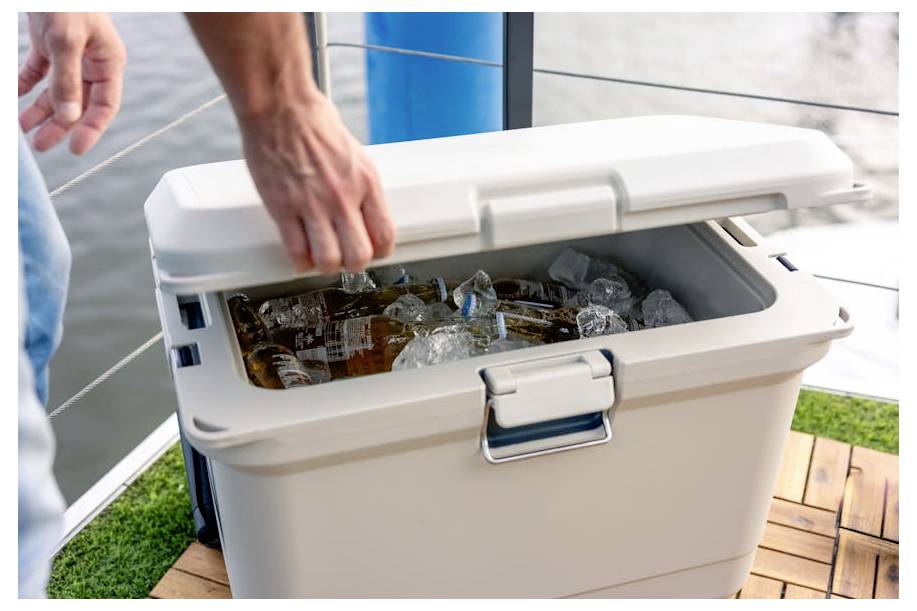 Een persoon opent een witte koelbox gevuld met ijs en flessen met dranken op een terras.