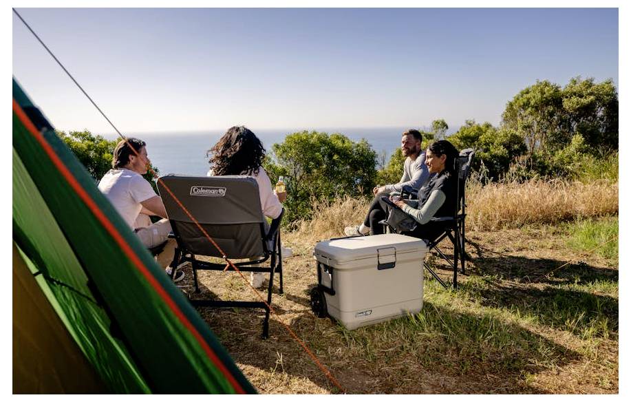 Vier mensen zitten in campingstoelen rond een koelbox op een schilderachtige kampeerplaats aan zee, met een tent op de voorgrond en uitzicht op de oceaan.