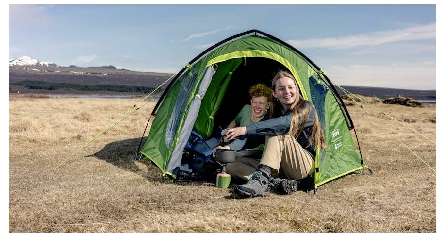 Twee mensen zitten in een groen kampeerttent op een droog grasterrein, glimlachend terwijl zij buiten eten of een drank bereiden.