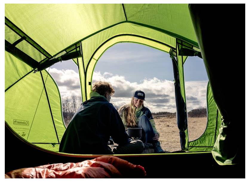 Twee mensen zitten buiten een groene campingtent te praten in een zonnig veld; een slaapzak is zichtbaar in de tent.