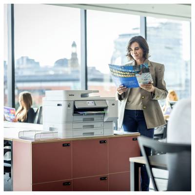 Vrouw die een brochure leest naast een Brother kantoorprinter in een helder kantoor met collega's op de achtergrond.