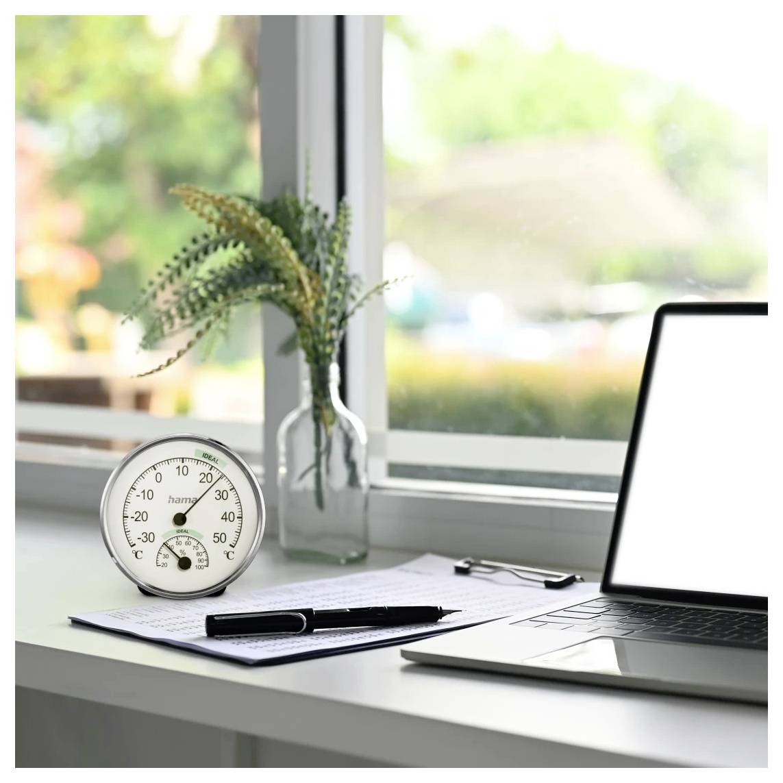Laptop op een bureau bij een raam met een papier, pen, klembord, thermometer en een vaas met bloemen in een helder thuiskantoor.