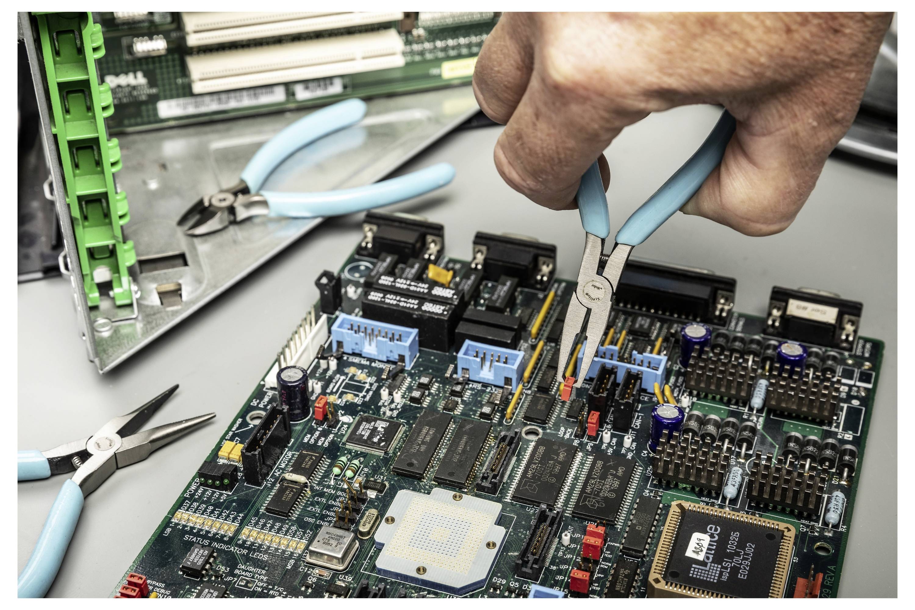 Hand die een naald-neus tang gebruikt om onderdelen op een printplaat aan te passen tijdens elektronicaherstellingen, met gereedschap en computeronderdelen in de buurt.