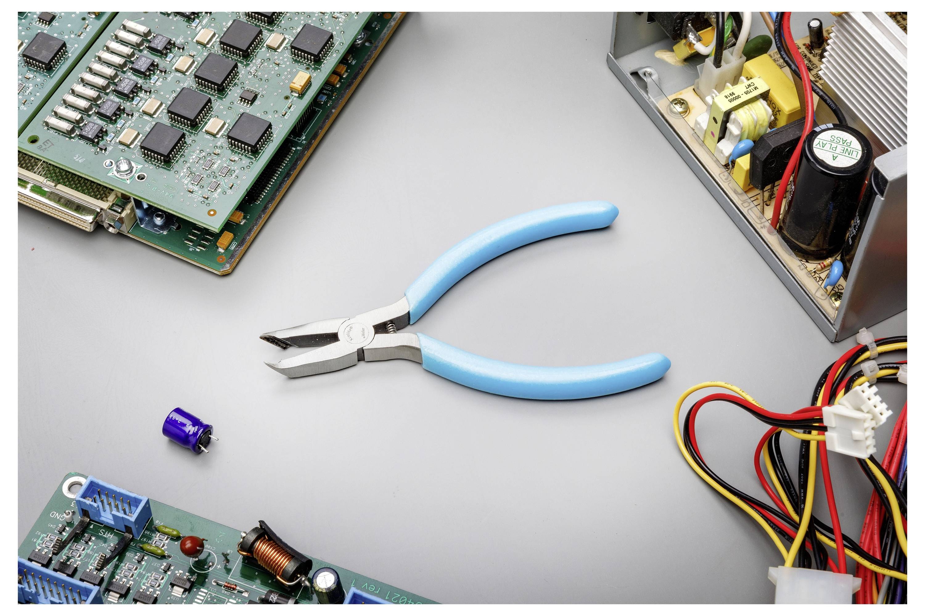 Blauwe tang gecentreerd op een werkbank met printplaten, draden en een open elektronisch apparaat, wat suggereert elektronica reparatie of montage.