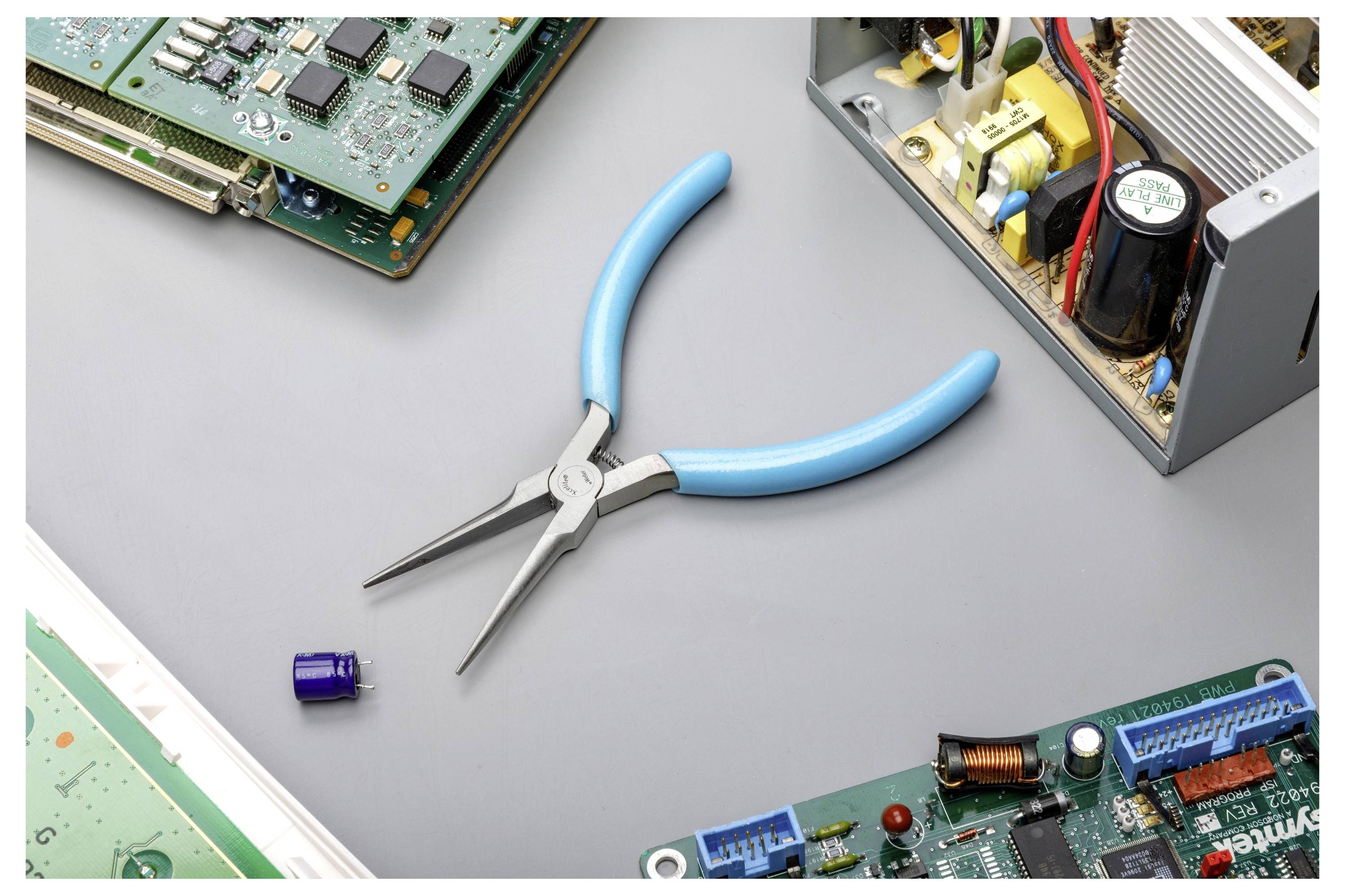 Blauwe buigtang gecentreerd op een grijs werkoppervlak, omgeven door elektronische printplaten en een open voedingscomponent.