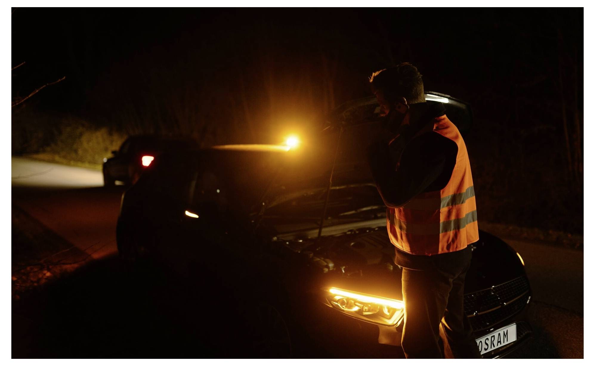 Een persoon in een reflectief vest inspecteert een kapotte auto 's nachts, verlicht door de koplampen van de auto en een amber waarschuwingslicht.