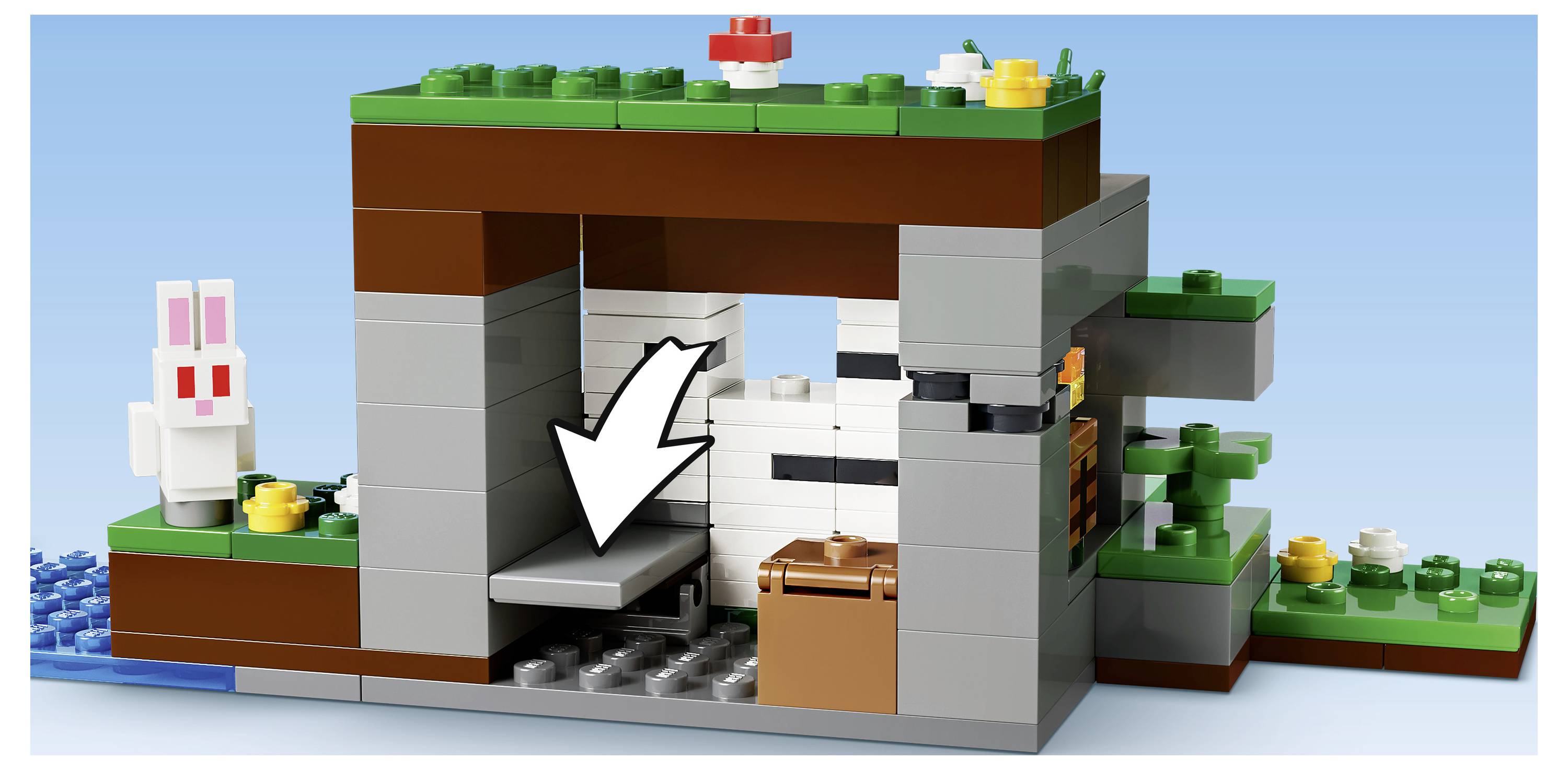 Een LEGO grot-ingangsscène met een witte pijl die naar een verborgen grijs platform binnenin wijst, naast een bruine kist en een wit konijnfiguur buiten.