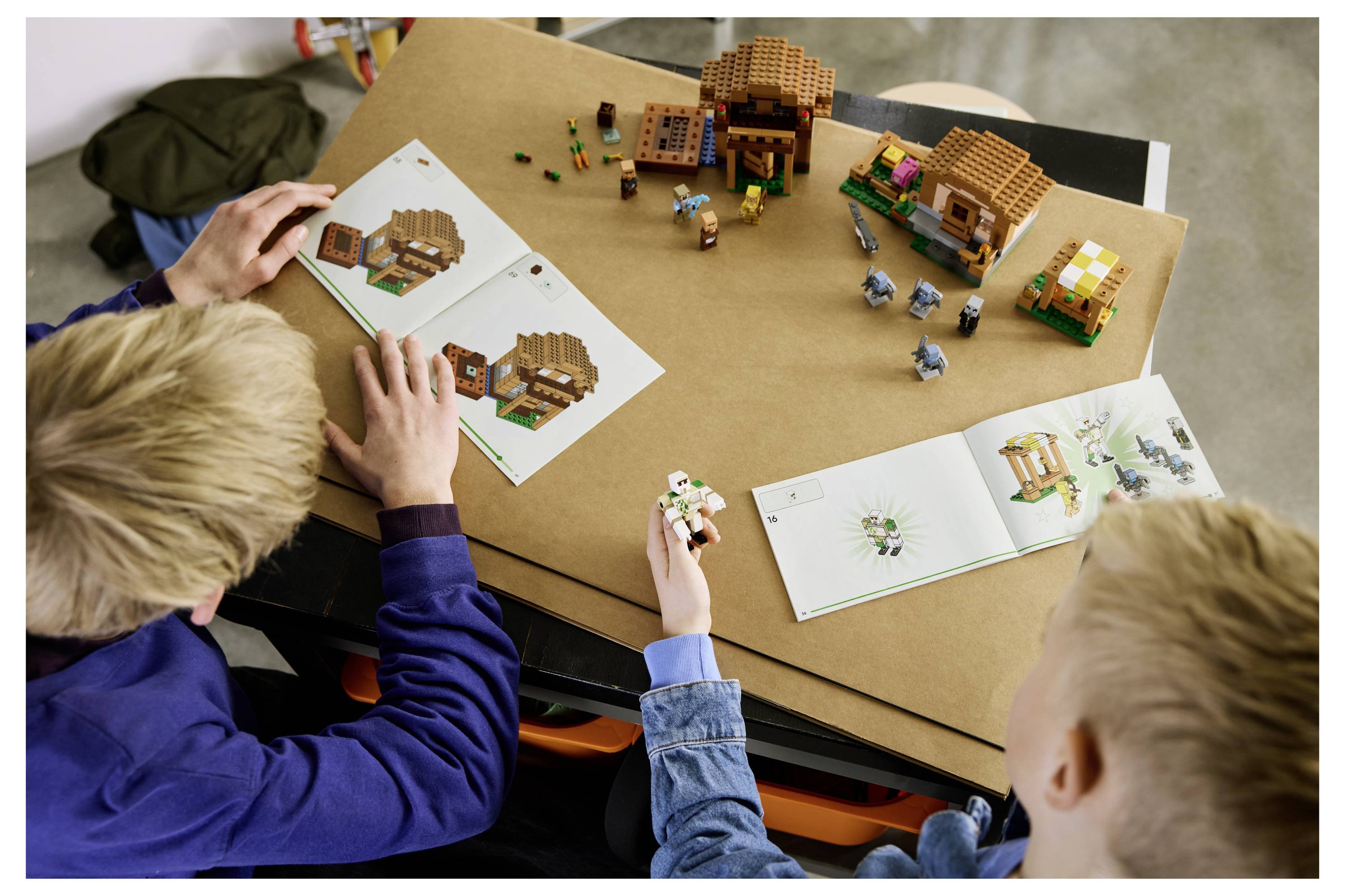 Twee kinderen bouwen een LEGO-dorp van instructieboekjes op een tafel, met kleine huisjes en figuren die op een kartonnen ondergrond zijn gerangschikt.