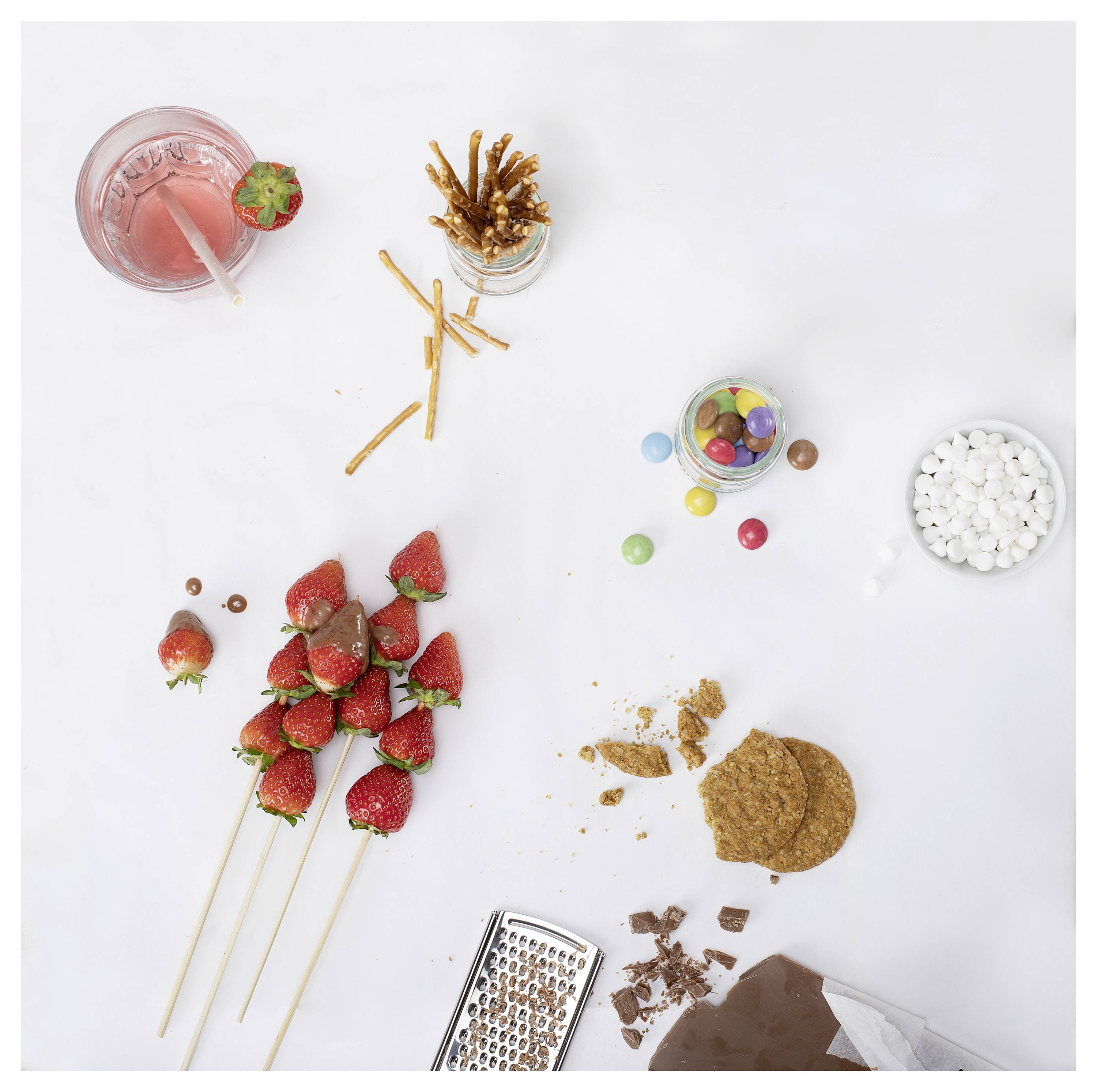 Bovenaanzicht van snoepgoed en snacks: aardbeiendrank, chocoladeovergoten pretzels, snoepkruik, marshmallows, aardbeienspiesjes, koekjes, geraspte chocolade en snoepstukjes op een witte achtergrond.