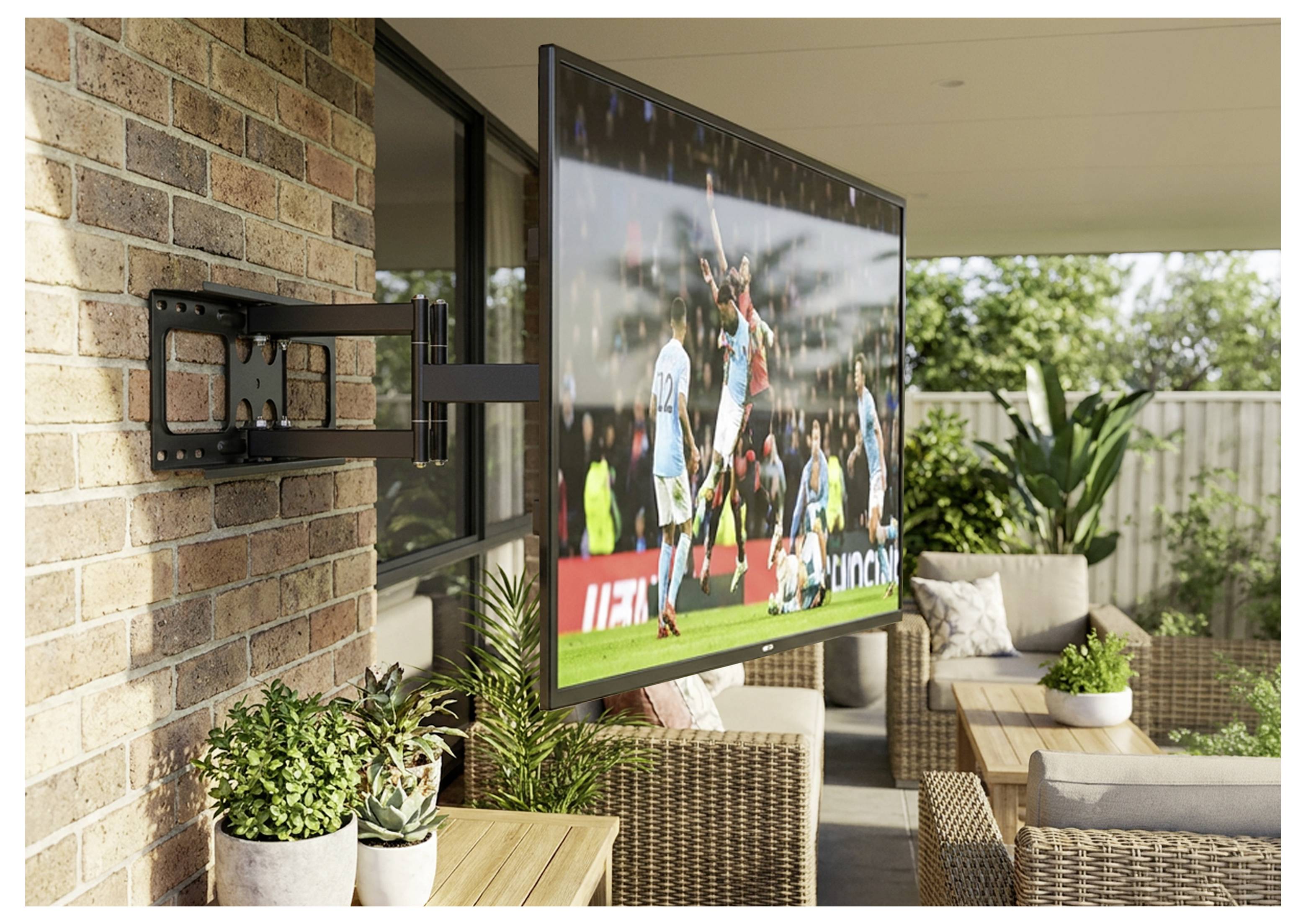 Flatscreen-tv gemonteerd op een bakstenen patiomuur, waarop een voetbalwedstrijd wordt weergegeven in een buitenzitgedeelte met rotan meubilair en planten.
